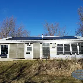 A modest single-story home outfitted with a rooftop solar panel system. The panels are neatly installed on the metal roof, optimizing the home for renewable energy. The front of the house features large windows and metal siding, with some sections covered for insulation or functional purposes. The property is set in a rural area with leafless trees and a clear blue sky.