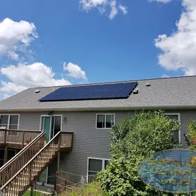 A two-story home with a rooftop solar panel array installed on the rear-facing roof. The house features a wooden staircase leading to an elevated deck overlooking a lush, green backyard. The solar panels are aligned in a single, compact section, complementing the house’s design. The scene is bright and sunny, with a vivid blue sky and scattered clouds.