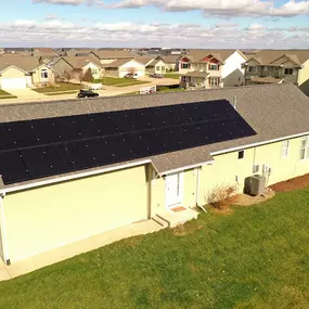 A single-story home in a suburban neighborhood equipped with a rooftop solar panel system. The solar array is installed on the rear slope of the roof, maximizing sunlight exposure. The house is surrounded by a neatly maintained lawn, and the neighborhood features rows of similar homes under a partly cloudy blue sky.