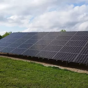 A ground-mounted solar panel array installed in a lush green field under a partly cloudy sky. The solar panels are arranged in a single, long row, tilted at an optimal angle to capture sunlight. The system is supported by a sturdy metal frame and positioned on a bed of gravel, blending seamlessly into the natural surroundings.