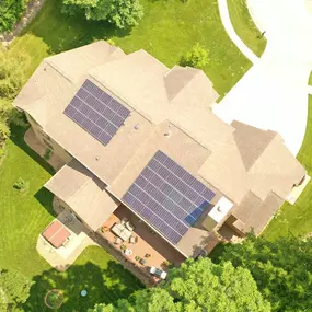 Aerial view of a residential home equipped with rooftop solar panels. The solar array is neatly installed on two sections of the roof, strategically placed to maximize exposure to sunlight. The house is surrounded by well-maintained landscaping, including lush green trees, a spacious lawn, and a paved driveway leading to a cul-de-sac.