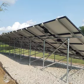 A rear view of a ground-mounted solar panel system showcasing the sturdy metal support structure. The solar panels are elevated on angled frames, supported by steel beams anchored on a gravel foundation. The image captures the underside of the array, revealing the precision engineering and durable design. The setup is surrounded by open green space under a bright, partly cloudy sky.