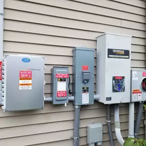 A wall-mounted setup of solar energy and backup power equipment installed on the exterior of a residential property. The system includes an Enphase™ energy management unit, a Generac™ transfer switch, disconnect switches, and a utility meter. Each component is neatly organized and labeled for safety and functionality. The setup is affixed to the siding of the home, with conduit pipes connecting the devices.