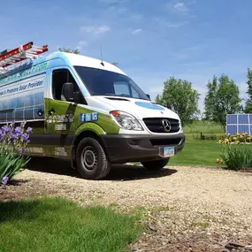 A branded company van for Energy Consultants Group parked on a gravel driveway, surrounded by vibrant purple flowers and a lush green landscape. The van features a custom wrap showcasing solar panels, the company logo, and promotional text, including 