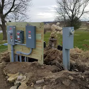 n outdoor solar power system installation featuring a wooden backing panel supporting electrical components such as disconnect switches and utility meter boxes. Adjacent to the wooden panel is a standalone utility meter mounted on a metal pole. The installation is set in a rural landscape, surrounded by a partially excavated area, open grassy fields, leafless trees, and a serene pond in the distance under a cloudy sky.