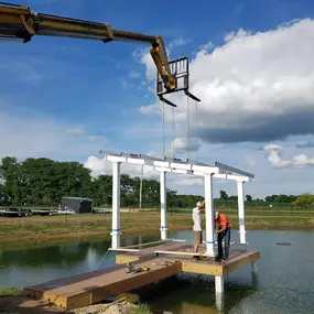 A solar panel mounting structure being installed over a pond, supported by white metal pillars anchored in the water. Two workers are on a wooden platform, preparing the structure while a crane arm hoists a component into position. The serene rural setting features a green landscape, a clear pond, and a partly cloudy blue sky.