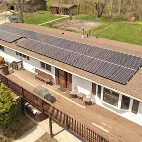 A rooftop solar panel system installed on a single-story home with a large wooden deck. The solar array spans nearly the entire length of the roof, showcasing a sleek and efficient design. The property is surrounded by trees, a small garden, and a propane tank, emphasizing a rural or semi-rural setting under a sunny sky.