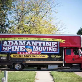 Adamantine Spine Moving truck parked in a driveway surrounded by greenery and a residential setting.