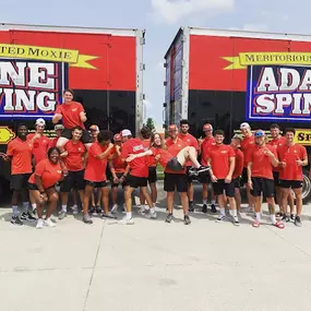 A large team of movers wearing red shirts posing in front of two Adamantine Spine Moving trucks.