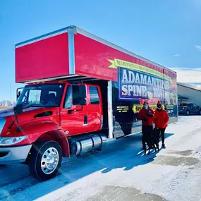 Adamantine Spine Moving truck parked on a snowy driveway with two employees and a dog in front.