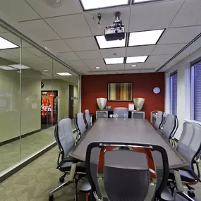 A modern conference room featuring a long rectangular table surrounded by ergonomic office chairs with mesh backs. The room has large windows on one side, providing a view of urban buildings, and a glass wall on the opposite side, creating an open and spacious feel. The red accent wall at the far end is decorated with contemporary art and flanked by decorative vases, adding a touch of elegance to the design. Ceiling-mounted lighting and a projector complete the setup.