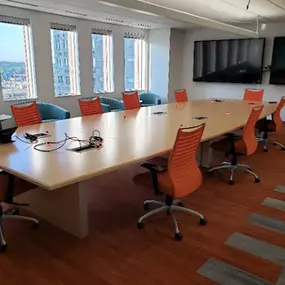 A bright and spacious conference room featuring a long, light-colored wooden table surrounded by modern orange office chairs with ergonomic designs. Large windows line one side of the room, offering a panoramic view of a cityscape and filling the space with natural light. The room also includes a wall-mounted flat-screen television for presentations, and the warm-toned hardwood flooring adds a welcoming touch.