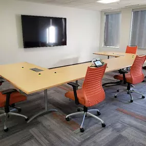 A small, modern meeting room featuring a unique, modular conference table with light wood surfaces and built-in connectivity ports. The table is surrounded by bright orange ergonomic chairs on wheels, providing both comfort and flexibility. The room includes a wall-mounted flat-screen television for presentations or video conferencing, with large windows on one side allowing natural light to enter.