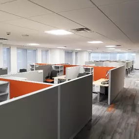 A modern office space featuring a grid of cubicles with gray walls and bright orange accent panels. Each cubicle is equipped with desks, shelving, and office chairs, providing a functional and organized workspace for employees. The space is illuminated by overhead lighting and natural light streaming in through large windows along the walls. The flooring features a contemporary gray and orange pattern, complementing the overall design.