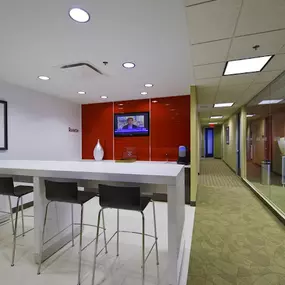 A stylish office lounge area featuring a sleek white high-top table with four modern black barstools. The lounge is accented by a bold red wall with a mounted TV displaying news and a decorative white vase adding a minimalist touch. Adjacent to the lounge, a glass wall offers a view into a conference room, and a carpeted hallway leads to additional office spaces. The neutral tones of the walls and flooring complement the vibrant red feature wall.