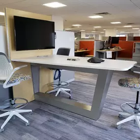 A collaborative workspace featuring a modern high-top table with a sleek white surface and a metallic base. The setup includes four adjustable swivel chairs with patterned seat cushions, adding a touch of color and comfort. A wall-mounted flat-screen TV is integrated into the design, perfect for presentations or video conferencing. The workspace is part of an open office layout, with cubicles and desks visible in the background.