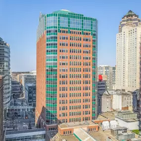 A vibrant cityscape featuring a tall, distinctive red-brick building with green-tinted windows and a modern geometric design. The building is surrounded by other high-rise structures of varying architectural styles, including classic brick facades and contemporary glass towers. The bright blue sky enhances the urban skyline, reflecting a clear, sunny day in a bustling downtown area.