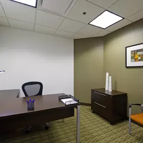 A compact and professional office space featuring a dark wooden desk with a sleek design, paired with a black mesh office chair. A bright orange guest chair adds a pop of color to the neutral-toned room, which includes olive-green and white walls for a calm yet modern aesthetic. A small cabinet with minimalist decor, including stacked white vases and framed abstract art, enhances the room's visual appeal.