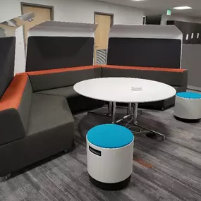 A modern and cozy office lounge area featuring a U-shaped sectional sofa with dark gray cushions and orange accents. The seating arrangement surrounds a round white table on a metallic base, creating a collaborative space for informal meetings or discussions. The setup includes two small, portable stools with white bases and bright blue cushions for additional seating. High-backed partitions provide privacy, and the area is set on contemporary gray and orange-patterned flooring.