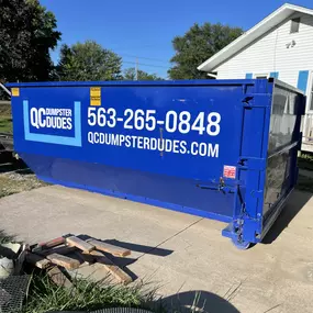 A blue QC Dumpster Dudes dumpster placed in a residential driveway, demonstrating its utility for home cleanouts and light construction waste.