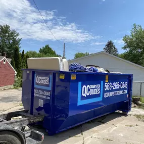 A blue QC Dumpster Dudes dumpster filled with furniture and miscellaneous items, located in a residential driveway.