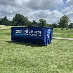 An empty blue QC Dumpster Dudes dumpster sits on a manicured green lawn, ideal for landscaping or outdoor cleanup projects.