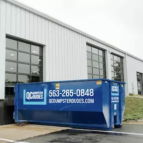 A large blue dumpster with QC Dumpster Dudes branding parked in front of a modern commercial building.