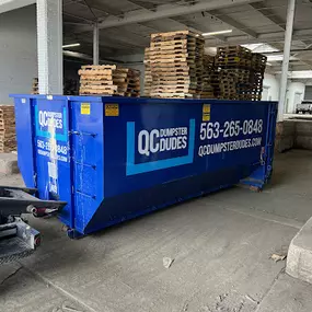 A QC Dumpster Dudes dumpster parked inside a warehouse filled with wooden pallets, ideal for industrial or commercial debris removal.