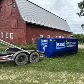 A blue QC Dumpster Dudes dumpster placed next to a red barn in a rural setting, perfect for agricultural or farm cleanups.