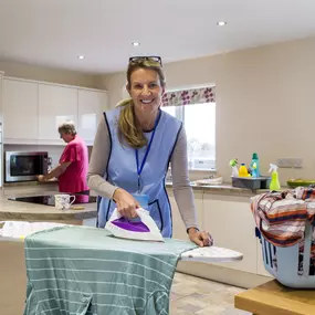 Care worker making a home visit. Female carer is ironing in the kitchen to help an elderly woman. The elderly woman is standing at the kitchen bench behind but not in focus.