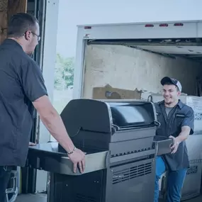 Two delivery personnel are moving a high-quality outdoor grill out of a truck. The background shows the inside of the delivery truck, with other appliances visible. The workers are wearing professional uniforms, highlighting their expertise and care in handling the equipment