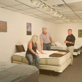 A retail showroom featuring several mattresses on display. A sales associate is assisting a couple who are testing the comfort of a mattress. The environment is clean, well-lit, and inviting, emphasizing personal attention and customer service.