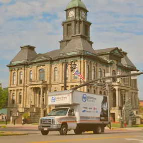 A branded delivery truck for Millersburg Electric is parked in front of a historic courthouse building. The truck showcases the company’s logo and appliance brands they carry. The photo conveys a sense of community presence and reliability.