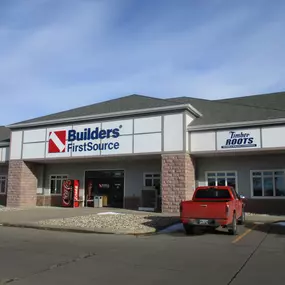 The exterior of the Builders FirstSource store, a modern building with a gray facade, white trim, and a bold Builders FirstSource sign prominently displayed. To the right is a sign for 