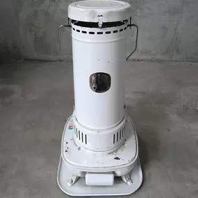 A vintage, white kerosene heater stands on a concrete floor, showcasing a simple yet practical design. The cylindrical heater features a small window for viewing the flame, and it sits atop a sturdy base with handles on either side for portability.