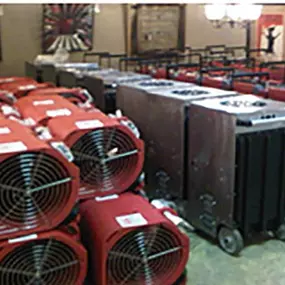 A storage room filled with professional-grade restoration equipment, including stacks of red air movers and industrial dehumidifiers on wheels. The equipment is neatly organized, ready for deployment in water damage restoration projects.