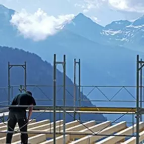 A construction worker assembles a wooden framework atop a scaffold with breathtaking mountain views in the background. The scene captures both the rugged natural beauty of Colorado and the precision of construction work.