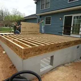 Two workers build a wooden deck frame attached to a residential home, creating the foundation for an outdoor living space. The frame sits on a concrete base, demonstrating the early stages of construction.