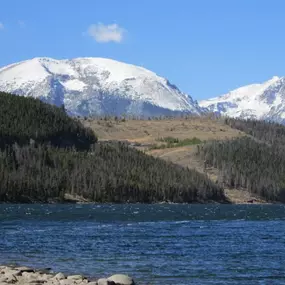 A serene view of a Colorado mountain range with a crystal-clear lake in the foreground. Snow-capped peaks rise in the distance, framed by dense forests and rocky terrain.