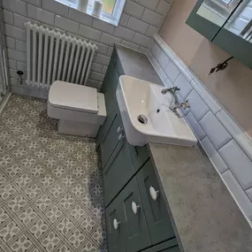 Compact bathroom renovation featuring a modern white sink, green cabinetry, patterned floor tiles, and a white radiator under a frosted window.