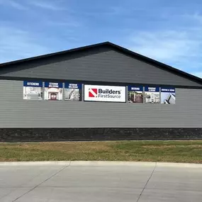 Side view of the Builders FirstSource building in Spencer, showcasing a modern structure with gray horizontal siding and a dark stone accent base. The wall is adorned with multiple promotional panels highlighting various services, such as kitchens, exterior materials, interior millwork, decking, and roofing & siding, all centered around the prominent Builders FirstSource logo. The neatly paved driveway in the foreground contrasts with the green grass lining the building, while a vivid blue sky w