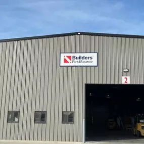 Exterior view of the Builders FirstSource warehouse in Spencer, featuring a large industrial building with a sleek gray metal siding and a gabled roof. The prominent Builders FirstSource logo is displayed at the top center of the building. Below, a wide open bay labeled with the red number '2' provides a glimpse into the warehouse interior, where forklifts and materials are visible. Smaller rectangular windows line the left side, and the bright blue sky with soft, wispy clouds creates a crisp an