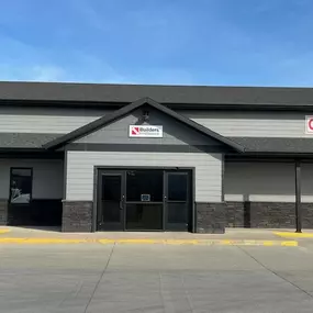 Exterior view of the Builders FirstSource store in Spencer, featuring a modern gray building facade with contrasting dark stone accents along the lower portion. The building has a clean, symmetrical design with large glass double doors at the center and bold red signage for 'Cabinets' on the right. The backdrop highlights a bright blue sky with no clouds, enhancing the fresh and inviting appearance of the store.