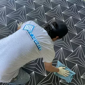 A craftsman cleaning up before grouting a black-and-white geometric cement tile floor, wearing a branded Davie Mac Tile shirt.