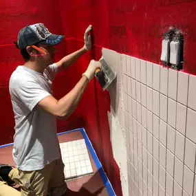 A craftsman applying white tiles to a red waterproofed wall in preparation for a shower installation.