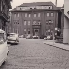 Eine historische Aufnahme des Rathauses II in Oldenburg mit Blick auf die gepflasterte Straße und parkende Autos aus vergangenen Jahrzehnten. Das Bild vermittelt den Charme des alten Stadtbildes.