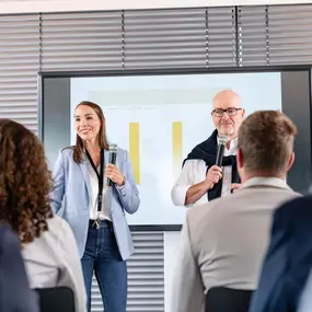 Ein professionell ausgestatteter Konferenzraum in der Alten Stadtkasse Oldenburg, der sich ideal für Schulungen, Vorträge und Präsentationen eignet. Auf dem Bild halten zwei Vortragende eine Präsentation vor einem aufmerksamen Publikum. Der Raum ist mit moderner Präsentationstechnik und komfortablen Sitzmöglichkeiten ausgestattet.