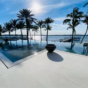 This expansive waterfront oasis showcases Epic Watershapes' expertise in creating luxurious and inviting outdoor living spaces. Our team designed and built this stunning pool area, featuring a sleek, geometric pool with a vanishing edge that seems to melt into the horizon. The expansive travertine deck provides ample space for lounging and entertaining, while carefully positioned palm trees offer shade and enhance the tropical ambiance.  A private dock extends into the serene water, inviting res