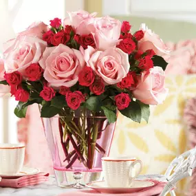 A stunning bouquet of pink roses and red spray roses elegantly arranged in a pink glass vase, placed on a cozy table setting with tea cups and dessert. The vibrant colors of the flowers beautifully contrast with the soft pastel background, creating a romantic and inviting atmosphere. Perfect for gifting on Valentine's Day, anniversaries, or as a thoughtful gesture to brighten someone’s day.