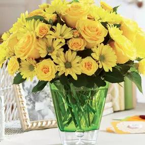 A cheerful arrangement of bright yellow roses and daisies displayed in a vibrant green glass vase, creating a sunny and uplifting centerpiece. The bouquet is set on a light and airy table with decorative accents, including a photo frame, wicker details, and a greeting card. This floral design is ideal for spring celebrations, birthdays, or sending a warm 'get well soon' message.